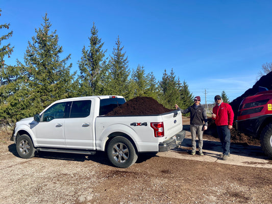 Two men smiling next to a white pickup truck loaded with premium dark brown mulch, with a large dark brown mulch pile and a red loader in the background. Buy bulk mulch at Raney Tree Care for quality landscaping supplies.