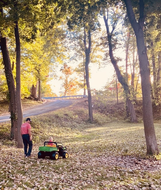 Dad in Red Raney Tree Care Shirt Walking Next To His Son Riding In A John Deere Mini Green Tractor In A Yard With Large Trees and Leaves on the Ground and Sunshine Peaking Through